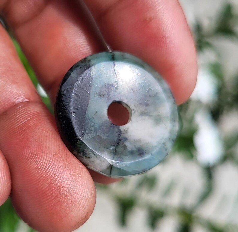 May include: A circular, donut-shaped pendant featuring a polished surface with a marbled pattern of deep forest green, charcoal gray, and creamy white tones. The piece has a smooth, rounded edge and a centered circular hole for threading.