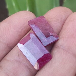 May include: Two rectangular, faceted ruby gemstones. The gems are a deep red colour with a polished surface, and are held in a person's hand. The gemstones have a rectangular shape with cut corners, and are displayed against a blurred green background.
