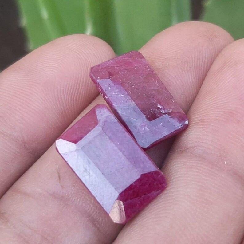 May include: Two rectangular, faceted ruby gemstones. The gems are a deep red colour with a polished surface, and are held in a person's hand. The gemstones have a rectangular shape with cut corners, and are displayed against a blurred green background.