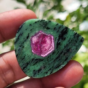 May include: A polished, teardrop-shaped ruby in zoisite gemstone featuring a vibrant, hexagonal magenta-pink ruby crystal embedded in the centre of a mottled green and black zoisite matrix with a smooth, flat surface and defined edges.