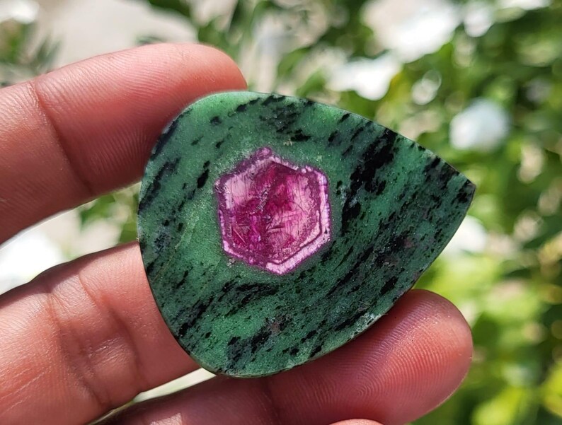 May include: A polished, teardrop-shaped ruby in zoisite gemstone featuring a vibrant, hexagonal magenta-pink ruby crystal embedded in the centre of a mottled green and black zoisite matrix with a smooth, flat surface and defined edges.