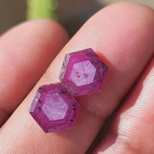 May include: Two hexagonal, faceted gemstones with a deep magenta hue. The gems have a complex internal structure and dark inclusions, held in a person's hand. The gems are likely rubies, showcasing a natural, unpolished appearance.