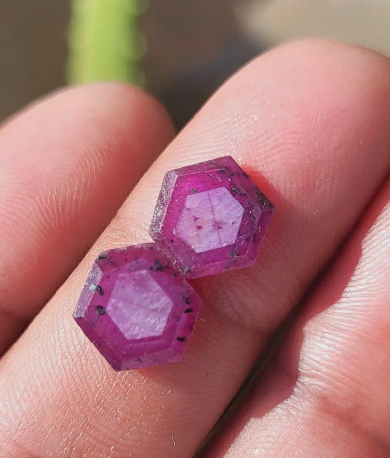 May include: Two hexagonal, faceted gemstones with a deep magenta hue. The gems have a complex internal structure and dark inclusions, held in a person's hand. The gems are likely rubies, showcasing a natural, unpolished appearance.