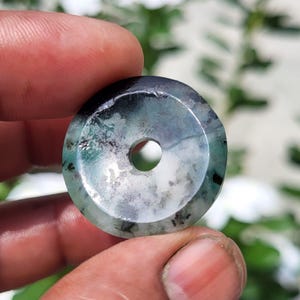 May include: A circular stone pendant featuring a polished surface with a mottled pattern of deep forest green, charcoal gray, and translucent white. The piece has a smooth, beveled edge and a small, centered circular hole for suspension.
