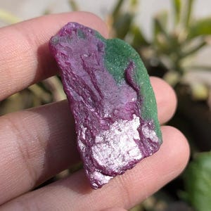 Peut inclure: Un spécimen minéral brut et non poli présentant une section magenta-violet vif à côté d'une section vert profond contrastante. La surface de la pierre affiche une texture cristalline naturelle avec des reflets métalliques brillants.