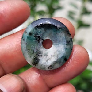 May include: A circular stone pendant featuring a polished surface with a mottled pattern of deep forest green, translucent white, and dark charcoal black inclusions, centered by a small, perfectly round hole for threading.