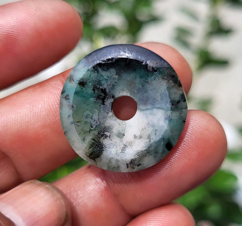 May include: A circular stone pendant featuring a polished surface with a mottled pattern of deep forest green, translucent white, and dark charcoal black inclusions, centered by a small, perfectly round hole for threading.