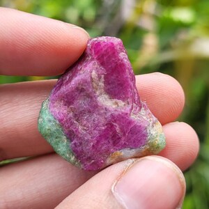 May include: A raw, unpolished gemstone featuring a vibrant, deep magenta-pink center with textured, crystalline surfaces. The base and edges of the stone display contrasting patches of opaque, pale green mineral material with a rough, natural finish.