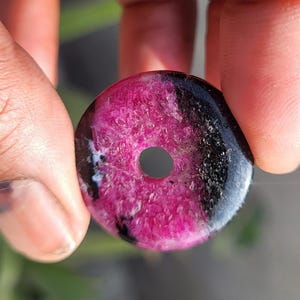 Può includere: Un ciondolo rotondo in pietra preziosa rosa e nera con un foro centrale. La sezione rosa ha un aspetto strutturato e cristallino, mentre le aree nere sono lisce e lucide. Il ciondolo è tenuto tra due dita.