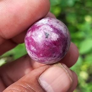 May include: A round, polished gemstone with a deep magenta hue, marbled with white and gray inclusions. The sphere is held between fingers, showcasing its smooth, reflective surface. The background is a blurred green.