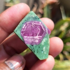May include: A natural gemstone with a striking magenta crystal set within a green matrix. The stone, held in a hand, has a geometric, hexagonal shape, highlighting its raw, unpolished form. The green matrix has black speckles, adding to its natural appeal.