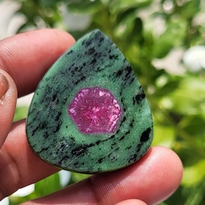 May include: A teardrop-shaped cabochon gemstone featuring a vibrant magenta hexagonal centre surrounded by a mottled green and black matrix. The stone displays a smooth, polished surface with distinct colour variations and natural mineral inclusions.