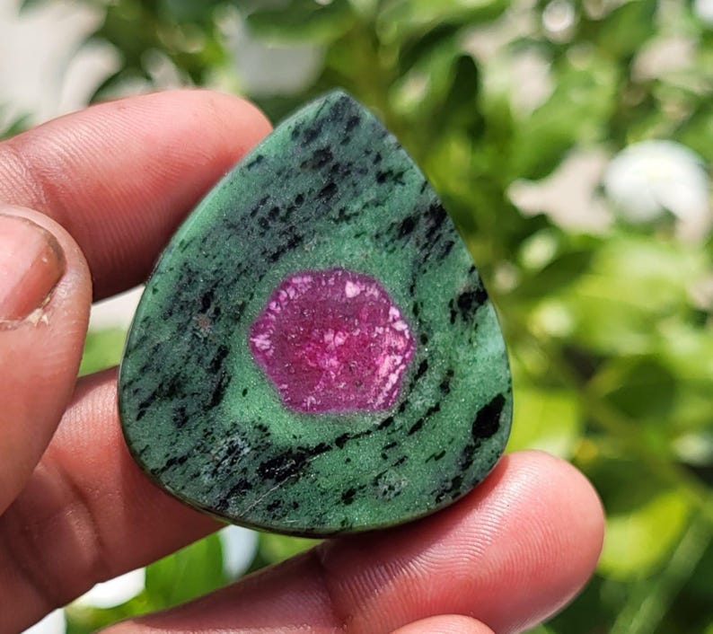 May include: A teardrop-shaped cabochon gemstone featuring a vibrant magenta hexagonal centre surrounded by a mottled green and black matrix. The stone displays a smooth, polished surface with distinct colour variations and natural mineral inclusions.