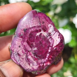 Puede incluir: Una piedra preciosa de rubí pulida y de corte plano que presenta una paleta de colores magenta intenso y carmesí. La piedra muestra un patrón de crecimiento interno hexagonal distintivo con fracturas superficiales visibles y un acabado brillante.