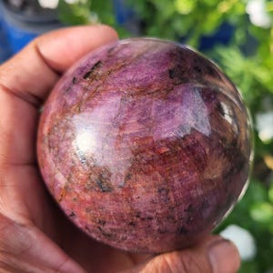 Può includere: Una pietra di rubino lucida e sferica caratterizzata da profonde tonalità magenta e viola con evidenti inclusioni scure e venature naturali su tutta la superficie, che presenta una consistenza liscia e riflettente e una forma solida.