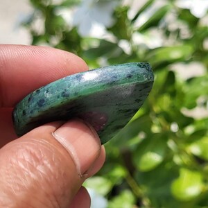 May include: A polished, teardrop-shaped gemstone featuring a deep green base with dark speckled inclusions and a distinct, smooth magenta-coloured patch visible on the surface. The stone has a rounded edge and a glossy, reflective finish.