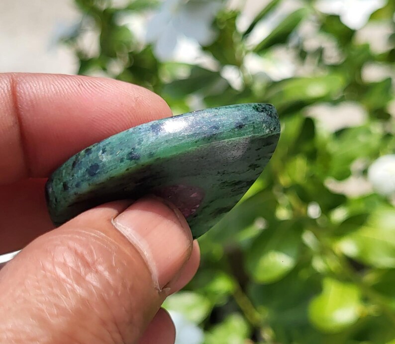 May include: A polished, teardrop-shaped gemstone featuring a deep green base with dark speckled inclusions and a distinct, smooth magenta-coloured patch visible on the surface. The stone has a rounded edge and a glossy, reflective finish.