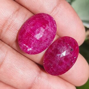 May include: Two oval-shaped, faceted rubies with a deep red hue. The gemstones are displayed against a neutral background, showcasing their polished surfaces and intricate cuts. These rubies are ideal for jewelry making.