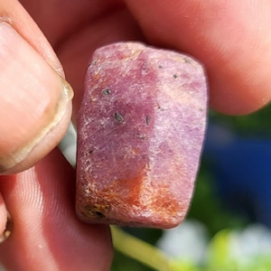 Könnte beinhalten: Ein roher, unpolierter Rubin-Edelstein mit rechteckiger, blockartiger Form. Der Stein zeigt einen tiefen rosa-roten Farbton mit sichtbaren natürlichen Einschlüssen, dunklen Mineralflecken und einer leicht durchscheinenden, texturierten Oberfläche.