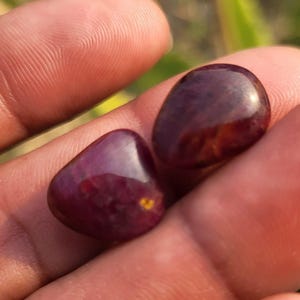 Puede incluir: Dos piedras preciosas pulidas, de color rojo intenso, con forma lisa y redondeada, se sostienen en una mano. Las piedras tienen un acabado brillante, que refleja la luz y muestra su rico color. Las gemas parecen ser piedras naturales.
