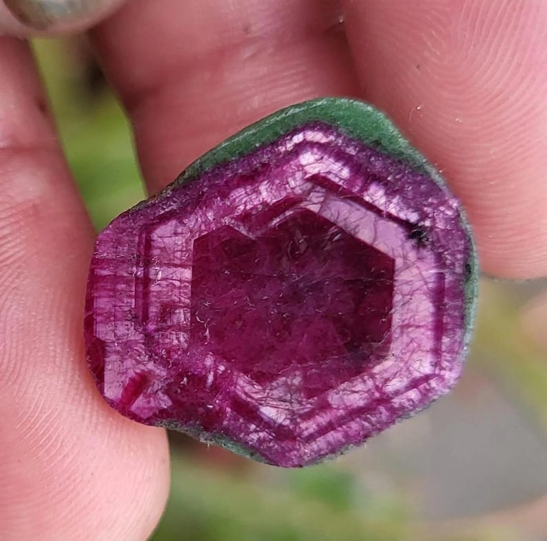 May include: A magenta-coloured gemstone with a green border. The stone is hexagonal, with visible internal structures and a polished surface. The gem's natural beauty is highlighted by its facets.