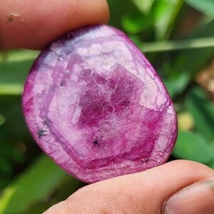 May include: A polished, oval gemstone in a deep magenta colour. The stone is translucent, with visible internal patterns and inclusions. The surface reflects light, creating a shimmering effect. The gemstone is held between two fingers.