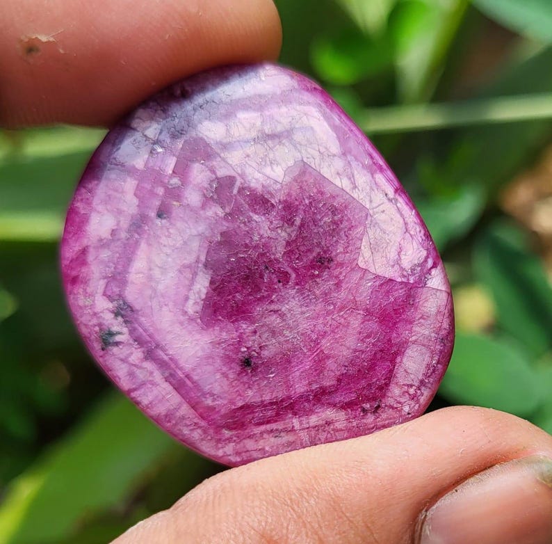 May include: A polished, oval gemstone in a deep magenta colour. The stone is translucent, with visible internal patterns and inclusions. The surface reflects light, creating a shimmering effect. The gemstone is held between two fingers.
