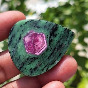 May include: A teardrop-shaped ruby zoisite gemstone featuring a vibrant magenta hexagonal crystal inclusion centred within a mottled green and black matrix. The polished stone displays a smooth surface with distinct colour variations and textures.