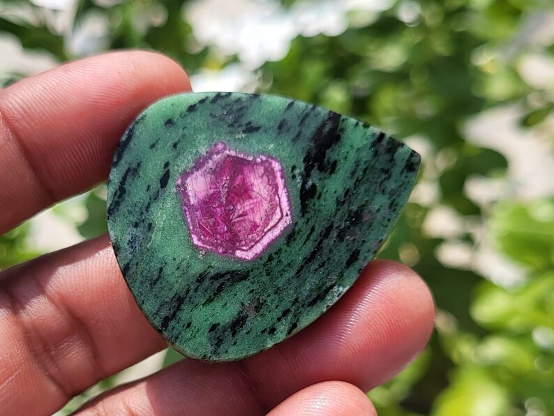 May include: A teardrop-shaped ruby zoisite gemstone featuring a vibrant magenta hexagonal crystal inclusion centred within a mottled green and black matrix. The polished stone displays a smooth surface with distinct colour variations and textures.