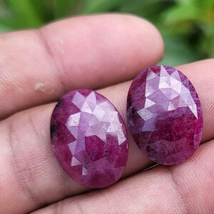 Puede incluir: Dos rubíes facetados de forma ovalada, de color rojo intenso. Las piedras preciosas se sostienen en una mano, mostrando sus superficies pulidas y cortes intrincados. Los rubíes reflejan la luz, resaltando su brillo.