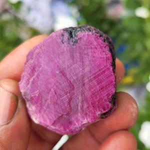 May include: A raw, unpolished ruby gemstone with a vibrant magenta-pink centre and dark, textured edges. The surface displays natural crystalline striations and a semi-translucent quality, showcasing the mineral's irregular, organic shape.