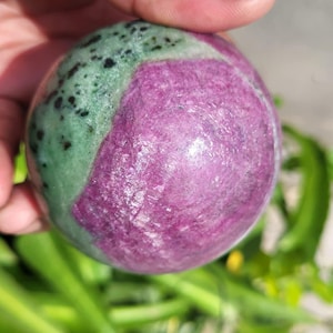 Może przedstawiać: Wypolerowana kula z rubinu w zoisycie. Kamień podzielony jest na dwie części: żywy odcień magenty i nakrapiany zielony obszar. Kula trzymana jest w dłoni, z zielonymi liśćmi w tle.