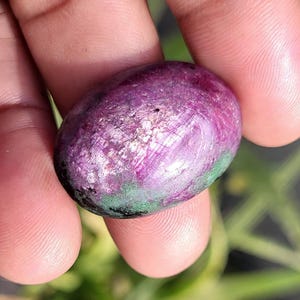 Op de afbeelding: Een gepolijste, ovaalvormige edelsteen met een mix van kleuren. De steen heeft een dieppaarse tint met roze en groene strepen. Het oppervlak heeft een glanzende afwerking, die licht reflecteert. De edelsteen wordt in een hand gehouden, met een vage groene achtergrond.