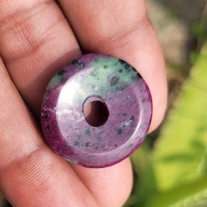 Può includere: Una perla rotonda e lucida di pietra preziosa con un foro al centro. La perla presenta una miscela di colori, tra cui rosso intenso, verde e nero. La pietra ha una finitura liscia e lucida e il foro è perfettamente centrato.