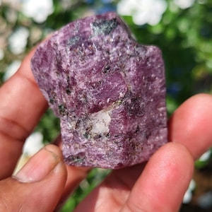 Puede incluir: Un espécimen de cristal de rubí en bruto y sin pulir que presenta un tono magenta y púrpura intenso con inclusiones minerales oscuras visibles y una pequeña mancha de matriz de cuarzo blanco incrustada en la superficie rugosa.