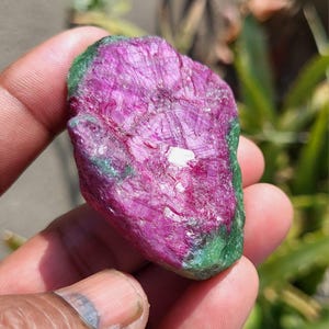 Peut inclure: Une pierre précieuse de rubis brute, d'un magenta vif, avec des touches de vert profond. La pierre naturelle présente une structure cristalline. Elle est tenue dans une main, mettant en valeur sa beauté brute.