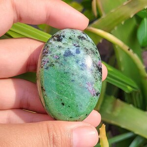 Op de afbeelding: Ovaalvormige gepolijste steen met een groene basiskleur, bespikkeld met zwart en vlekjes roze en rood. De steen wordt in een hand gehouden, met groen gebladerte op de achtergrond.