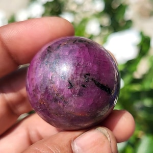 Op de afbeelding: Een gepolijste, bolvormige edelsteen met een diepe magenta en paarse tint met zichtbare donkere zwarte minerale insluitingen over het hele oppervlak. De steen heeft een gladde, reflecterende textuur en vertoont natuurlijke kleurvariaties.