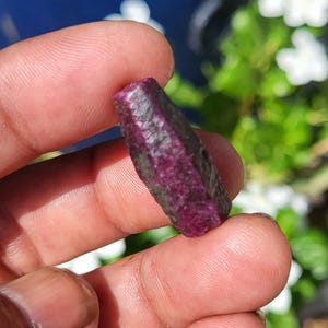 May include: A raw, hexagonal ruby crystal with a deep magenta and dark purple colour palette. The gemstone features a rough, unpolished surface texture with visible natural facets and a cylindrical, elongated shape held between two fingers.