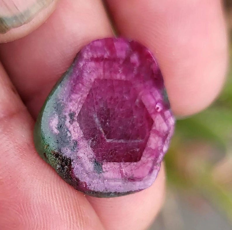 May include: A raw gemstone in shades of magenta and green. The stone has a hexagonal pattern in the center. The edges are dark green and black. The overall shape is irregular and organic. The stone is translucent.