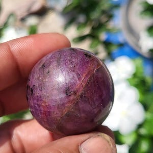 Op de afbeelding: Een gepolijste, bolvormige robijn met een diepe magenta en paarse tint, voorzien van zichtbare natuurlijke insluitingen, donkere mineraalvlekken en een duidelijke diagonale breuklijn over het gladde, afgeronde oppervlak van de steen.