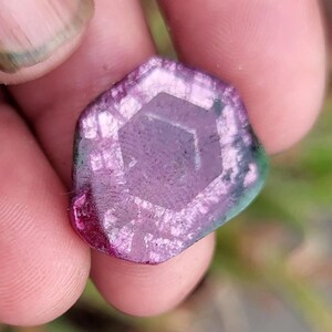 May include: A multi-faceted gemstone with a hexagonal pattern at its core. The stone displays a colour gradient, shifting from deep magenta to lighter purple and green tones. The gem is held between fingers.