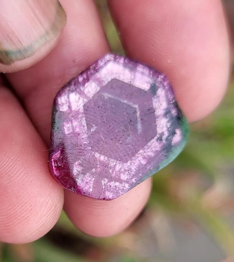 May include: A multi-faceted gemstone with a hexagonal pattern at its core. The stone displays a colour gradient, shifting from deep magenta to lighter purple and green tones. The gem is held between fingers.