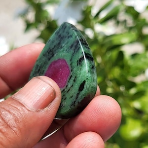 May include: A polished, teardrop-shaped zoisite gemstone featuring a vibrant, solid magenta-pink centre surrounded by a mottled green and black matrix. The stone has a smooth, reflective surface and a distinct, organic pattern throughout the mineral.