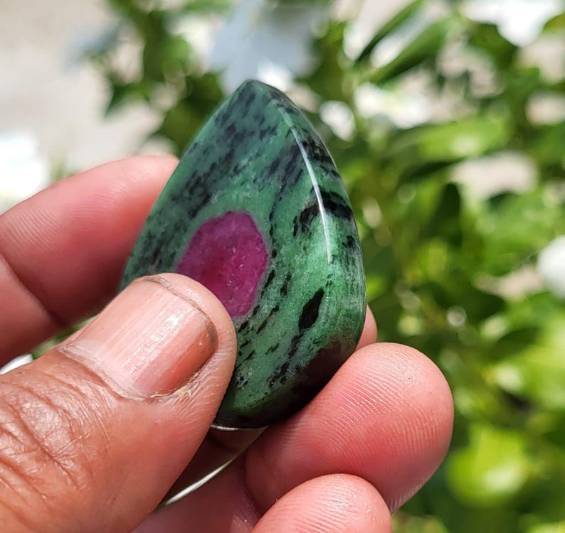 May include: A polished, teardrop-shaped zoisite gemstone featuring a vibrant, solid magenta-pink centre surrounded by a mottled green and black matrix. The stone has a smooth, reflective surface and a distinct, organic pattern throughout the mineral.