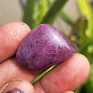 Puede incluir: Una piedra preciosa pulida y lisa, de color magenta intenso, con sutiles vetas internas y pequeñas inclusiones oscuras. La piedra se sostiene en una mano, con un fondo verde borroso.
