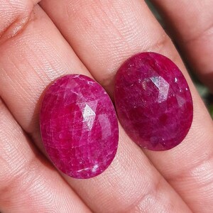 May include: Two oval-shaped, faceted rubies with a deep red hue. The gemstones are held in a hand, showcasing their polished surfaces and intricate cuts. The rubies are a vibrant, eye-catching color.