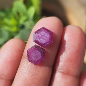 May include: Two faceted, hexagonal gemstones with a deep magenta hue. The gems are held between fingers, showcasing their geometric cut and natural color. The background is blurred, highlighting the gemstones.