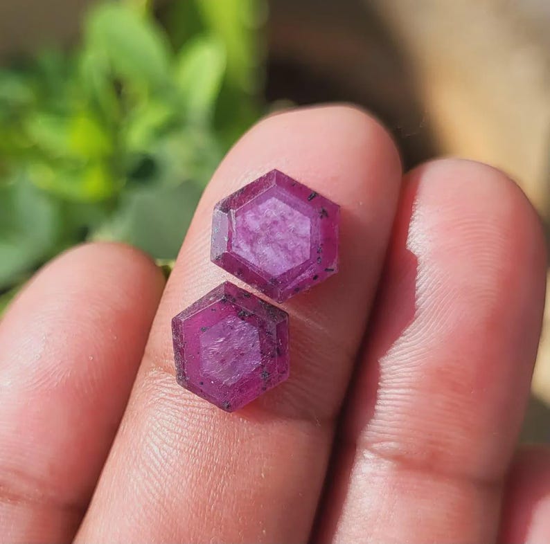 May include: Two faceted, hexagonal gemstones with a deep magenta hue. The gems are held between fingers, showcasing their geometric cut and natural color. The background is blurred, highlighting the gemstones.