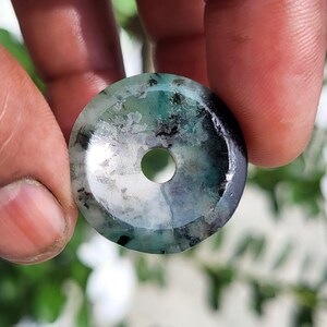 May include: A circular, flat pendant with a central hole, featuring a polished surface with a mix of translucent green, white, and dark grey inclusions. The stone displays natural mineral patterns and a smooth, reflective finish throughout.